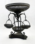 Fruteira de mesa em metal, Art nouveau de estrutura elevada, composta por bandeja circular superior e três pequenos cestos suspensos articulados. Peças vazadas com decoração geométrica em grega, sustentadas por armação em metal com elementos curvilíneos. Apresenta sinais de oxidação e desgaste compatíveis com o tempo. Alt.: 29 cm. 