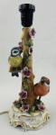 Abajur em porcelana, de inspiração naturalista, com tronco de árvore em relevo, flores e duas aves modeladas e policromadas. Base de formato recortado, com decoração em pintura dourada e detalhes florais. Montagem para iluminação elétrica, com soquete e fiação. Inscrição manuscrita na parte inferior. Alt.: 31 cm. Apresenta bicados nas flores e restauro no rabo de um pássaro. 