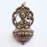Pequena pia de água benta de parede em metal representando o Apóstolo Santiago. Apresenta o busto de São Tiago com auréola, cajado e cabeça de peregrino emoldurado por guirlanda, concha decorada com asas de querubins. Med 10,5 x 5,5 cm.