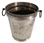 Antigo balde de gelo em metal espessurado a prata, borda decorada com ramagens e volutas em relevo, duas alças laterais móveis em argola e base circular moldurada. Corpo com dedicatória gravada alusiva à Diretoria de Aeronáutica do Galeão, datada de 1961/1966. Apresenta oxidação, riscos e desgastes. Medindo: 21 x 18 cm.