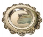 Pequena bandeja circular em metal espessurado a prata, centro liso e rebaixado, aba larga com borda recortada, ornamentada com concheados e volutas em relevo, de inspiração Rococó. Apresenta riscos superficiais e marcas do tempo. Medindo: 14 cm de diâmetro.