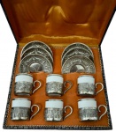 WOLFF. Conjunto de seis xícaras de café em porcelana branca com filetes prateados, acondicionadas em suportes vazados com alças curvas, acompanhadas de seis pires em metal espessurado a prata, decorados com motivos florais e caneluras em relevo. Em estojo forrado de cetim alaranjado, com identificação da marca. Medidas da caixa: 7,5 x 27,5 x 26 cm.