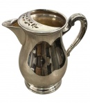 Antiga jarra em metal espessurado a prata, com corpo bojudo liso, base circular moldurada, bico saliente e alça curva com detalhes em relevo. Abertura parcialmente coberta por aparador de gelo decorado com motivos vegetalistas vazados. Medidas: 22 x 21 x 12 cm.