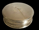 Pequena caixa circular em metal espessurado a prata, com tampa removível de superfície texturizada e monograma `IHS` envolto por raios gravados. Corpo decorado com frisos de volutas em relevo. Apresenta riscos e desgaste. Medidas: 4 cm de altura x 9,3 cm de diâmetro.