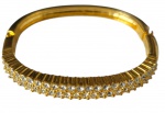 Bracelete em metal dourado, de aro rígido e formato oval, articulado por dobradiça e com fecho de encaixe. Frente ornamentada com duas fileiras de pedras incolores facetadas e frisos transversais em relevo; parte posterior lisa e polida.