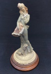 Escultura em resina policromada, representando dama com chapéu e vestido longo, acompanhada de cão, sobre base circular e pedestal de madeira. Apresenta marcas de uso e desgaste na pintura. Medidas: 26 x 14 cm.