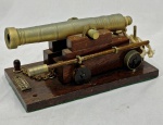 Miniatura de canhão naval em  bonze e metal dourado, sobre carreta de madeira com quatro rodas, acompanhada de vareta, balde, cordame e corrente. Base retangular de madeira com placa CARRONADA SÉC. XIX. Apresenta oxidação no metal e desgaste no acabamento da madeira. Medidas: 8 x 19 x 10 cm.