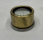 Peça com corpo cilíndrico em metal dourado, lente convexa e flange circular para fixação.  Medidas: 30 x 30 cm.