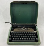 Antiga máquina de escrever manual portátil, com estrutura metálica esmaltada em verde, teclado preto e mecanismo de tipos aparente. Apresenta oxidação nas partes metálicas, desgaste na pintura e marcas do tempo.