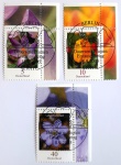 Alemanha, 2005  Lote com 3 selos  pertencentes à aclamada série regular "Blumen" (Flores), trazendo os valores de 5 Centavos ilustrando o delicado Açafrão (Michel: #2480), de 10 Centavos destacando a elegante Tulipa (Michel: #2484) e de 40 Centavos apresentando a charmosa Hepática (Michel: #2485). O design botânico em alta definição exibe as espécies em cores vibrantes contra fundos artísticos e esfumaçados. Todos os três exemplares deste belíssimo lote destacam-se por preservar a ampla margem superior da folha com as respectivas extensões do padrão floral e ostentam o cobiçado carimbo oficial de primeiro dia de circulação da série (Dauerserie Blumen Erstausgabetag Berlin), datados de 11.08.2005 (valor de 5c) e 08.09.2005 (valores de 10c e 40c), exibindo o contorno estilizado de uma flor como elemento gráfico do cancelamento