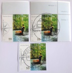 Alemanha, 2005  Lote com 3 selos comemorativos em celebração à tradicional profissão do carteiro fluvial na região de Spreewald (Michel: #2481). O belíssimo design de 55 Centavos retrata o carteiro conduzindo sua tradicional embarcação pelos canais cercados por uma densa vegetação verdejante, trazendo na lateral uma rosa dos ventos estilizada. Este lote oferece um excelente conjunto de estudo filatélico reunindo três variantes e estados de conservação distintos: um exemplar novo (MINT/MNH) preservando a margem esquerda com o desenho da rosa dos ventos completo; um exemplar em canto de folha superior direito, exibindo os nomes de localidades na borda (Wriezen, Letschin, Seelow) e cancelado com o cobiçado carimbo oficial de primeiro dia de circulação (Berlim, 11.08.2005) ilustrado com uma caixa de correio antiga; e um exemplar legitimamente circulado sobre fragmento, trazendo o carimbo postal de um centro de triagem regional (Briefzentrum)