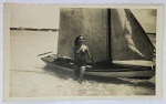 Foto Antiga Época (c. 1950) Jovem em Traje de Banho com Barco a Vela  Borda Canoa: Charmoso registro fotográfico em preto e branco que captura o estilo de vida e o lazer náutico de época. A imagem mostra uma jovem vestindo um maiô clássico, posando na água ao lado de um pequeno barco a vela de madeira em uma praia ou represa.