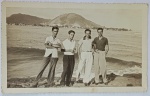 Foto Antiga Época (c. 1950) Jovens na Praia com Morro ao Fundo  Borda CanoaBelo registro em preto e branco retratando quatro jovens rapazes posando à beira-mar (provavelmente sobre rochas ou na faixa de areia). A imagem destaca a moda masculina casual da época, com camisas de gola polo, camisas de botão de manga curta e calças de alfaiataria de corte reto com passadores e cintos bem marcados. Ao fundo, vê-se a linha costeira com construções baixas e uma proeminente elevação montanhosa à beira-mar (característica de praias do litoral paulista ou fluminense, como a região de Santos/São Vicente ou Rio de Janeiro).