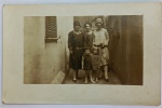 Foto Bilhete Postal Original (1928) Retrato Feminino e Infantil  Moda Anos 20 Charmoso e elegante registro social familiar focado no universo feminino da década de 1920. A imagem mostra três mulheres e duas crianças pequenas posando em um corredor ou entrada residencial de época, com uma janela de veneziana de madeira clássica ao fundo. O grande destaque colecionável são os trajes típicos dos "Anos Loucos" (anos 20), evidenciados pelos vestidos de corte reto com cintura baixa e, principalmente, pelos marcantes chapéus estilo cloché usados pelas mulheres.