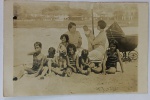 Foto Histórica (1928) Família no Santos Palace Hotel  Praia do José Menino, Santos - SPRegistro em tom sépia mostrando um grupo familiar sob um guarda-sol na praia, com destaque para a moda balnear infantil listrada dos anos 1920 e um raro carrinho de bebê antigo com capota e grandes rodas de raios.