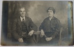 Foto Cartolina Postale Época (c. 1910/1920) Retrato de Casal com Relógio de Bolso  Papel Ubermor Solene e austero retrato de estúdio em tons de sépia e cinza-escuro capturando um casal de senhores de meia-idade posando assentados ao lado de uma mesa decorativa. A imagem é riquíssima em acessórios de época de grande apelo colecionável: Acessório Masculino: O cavalheiro veste terno completo de três peças (paletó, colete e calça), destacando uma imponente corrente de relógio de bolso (chatelaine) atravessando o colete. Acessório Feminino: A senhora, elegantemente trajada com blusa escura de gola estruturada e colar com pingente, segura firmemente em uma das mãos uma sombrinha ou guarda-sol fechado de época.