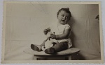 Foto Época (c. 1940/1950) Bebê Sentado em Banquinho com Bichinho de Pelúcia Antigo  Borda Canoa Meigo e expressivo retrato em preto e branco capturando um bebê sentado no topo de um pequeno banco redondo de madeira, posando em frente a um cenário de tecido improvisado. O bebê segura um bichinho de pelúcia antigo (formato de gatinho ou cachorrinho) com um lacinho no pescoço. No canto superior direito da imagem, nota-se discretamente a mão de um adulto dando suporte à cena de estúdio doméstico.