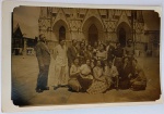 Foto Época (c. 1950) Grande Grupo de Turistas na Fachada de Catedral em Estilo NeogóticoO grupo está posicionado no pátio ou praça em frente a uma monumental igreja de arquitetura neogótica. A imagem destaca com grande riqueza de detalhes os portais ogivais (arcos apontados) de entrada, os frontões decorados, as colunetas e nichos esculpidos na pedra, além de janelas em arco gótico ao fundo, servindo como um excelente registro documental de patrimônio religioso do período
