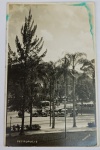 Foto Postal Antigo (c. 1930) Praça e Carros de Época em Petrópolis - RJ  Assinado R. Haack  Raro e belíssimo registro urbano em tom sépia da cidade imperial de Petrópolis - RJ. A composição destaca palmeiras e pinheiros em primeiro plano, uma mureta de canalização e uma fileira de automóveis antigos das décadas de 1920 e 1930 estacionados na avenida ao fundo, além da silhueta de casarios históricos e montanhas da região. Identificação e Assinatura no Negativo: Na base esquerda da imagem consta a identificação geográfica "PETROPOLIS" e, no canto inferior direito, a assinatura do renomado fotógrafo/editor de época: "R. HAACK".