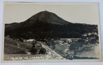 Foto Postal Antigo (c. 1940) Morro de Caxambu e Linha Férrea, Caxambu - MG Belíssimo e amplo registro panorâmico destacando o imponente Morro de Caxambu ao fundo. Em primeiro plano, a composição revela uma linha férrea (trilhos de trem) serpenteando a encosta, um pequeno vilarejo com casarios rústicos de época e a densa vegetação local.