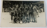 Foto Histórica Datada (1949) Time de Vôlei Militar do Quartel General de Recife - PE  Borda Canoa Espetacular e raro registro esportivo em preto e branco. A imagem captura um time de voleibol masculino composto por sete atletas posando em uma quadra aberta ao lado de seu treinador ou oficial uniformizado (vestindo quepe e farda militar). Os atletas trajam uniformes idênticos de regata com o emblema de uma ave heráldica estilizada (remetendo à Aeronáutica ou Infantaria) estampado no peito. Um dos jogadores agachados segura a bola clássica de vôlei de gomos brancos de época. Ao fundo, nota-se a rede, coqueiros e um quiosque/guarita do quartel.