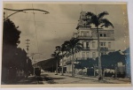 Raro Foto Postal Datado (1929) Grande Hotel de Guarujá com Bonde Elétrico  Endereçado a Santos  Espetacular e raríssimo registro que documenta a era de ouro da hotelaria e do transporte na Baixada Santista. A imagem destaca a suntuosa arquitetura eclética com torreões do lendário Grande Hotel de Guarujá (inaugurado no final do século XIX e demolido na década de 1960). À esquerda, a composição captura a infraestrutura pioneira de transporte da ilha, mostrando a fiação aérea, postes decorativos e um bonde elétrico clássico circulando sobre os trilhos na avenida arborizada com palmeiras.