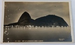 Foto Postal Noturna (c. 1950/1960) Pão de Açúcar e Urca Iluminados  Foto Postal ColomboNo topo de ambos os morros, veem-se as luzes acesas das estações do bondinho e, estendendo-se entre as duas montanhas, destaca-se o traço luminoso formado pelos cabos do teleférico iluminados na noite carioca. Na base da Urca, nota-se também um letreiro luminoso comercial típico do período