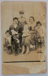 Foto Época (c. 1920/1930) Retrato de Família com Jovem Militar e Rica Dedicatória à Comadre Altiva Solene e expressivo retrato de estúdio de um grupo familiar composto por seis pessoas (pai, mãe, um jovem militar, duas meninas pequenas com grandes laços no cabelo e um bebê de colo vestindo touca clara). Destaque de Indumentária Militar: Ao fundo, em posição central, destaca-se um jovem em pé vestindo farda militar de época completa, incluindo túnica abotoada com ombreiras estruturadas e quepe rígido colonial (comum em corporações estaduais, forças públicas ou exército nas primeiras décadas do século XX).
