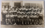 Foto Histórica Época (c. 1950/1960) Retrato Escolar de Turma Masculina com Professora Solene e tradicional retrato oficial de turma escolar de meados do século XX. A imagem captura um numeroso grupo composto por 28 alunos meninos (crianças e pré-adolescentes) elegantemente alinhados em três fileiras (sentados, agachados e em pé) na área externa da escola.