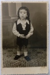 Foto Época (c. 1950) Retrato de Menino Nilson com Traje de Alfaiataria Infantil  Estúdio VAM  Borda CanoaA criança veste um refinado traje de alfaiataria infantil composto por colete escuro, shorts curto combinando, camisa de botão clara com gola estruturada, meias brancas de cano médio e sapatinhos sociais escuros. Ao fundo, nota-se o cenário cenográfico de estúdio com a base de uma coluna clássica.