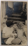 Foto Época (c. 1940/1950) Duas Jovem Mulheres com Bebê em Pátio Residencial  Papel Telado Fosco Meigo, íntimo e muito expressivo retrato familiar em plano médio. A imagem captura duas jovens mulheres assentadas no chão de um pátio residencial, olhando com extremo afeto e atenção para um pequeno bebê de colo envolto em um cueiro claro, que é segurado carinhosamente nos braços da mulher à esquerda.