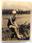Foto Época (c. 1940) Jovem de Maiô em Antigo Trapiche / Ruínas de Píer de MadeiraRaríssimo, poético e intrigante registro ao ar livre em tom sépia. A cena captura uma jovem mulher sorridente assentada sobre o que parece ser os destroços de madeira e metal de uma antiga embarcação, píer desativado ou estrutura de contenção náutica à beira-mar ou de um rio.