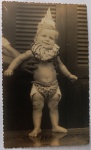Fotografia Vintage de Época  Bebê Fantasiado de Palhaço / Carnaval (c. 1950)Divertida e expressiva fotografia original de época (formato cartão com bordas recortadas em estilo ziguezague "guilhotina"). A imagem registra um bebê sorridente fantasiado, vestindo uma gola bufante franzida, um chapéu cônico pontudo (estilo Pierrô ou palhaço de Carnaval) e fralda decorada, sendo amparado pelas mãos de um adulto ao fundo. O clique captura uma autêntica cena doméstica de celebração ou mesversário de meados do século XX.