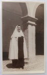 Fotografia Histórica (1961)  Tomada de Hábito na Ordem Terceira do Carmo (Marechal Deodoro - ALBela e solene fotografia original de época capturada em formato vertical. A imagem retrata uma religiosa (freira) posando de corpo inteiro em um ambiente de arquitetura sacra clássica, possivelmente o claustro ou pátio interno de um convento, mosteiro ou colégio religioso. A composição destaca os arcos estruturais de pedra e o piso decorado em ladrilhos hidráulicos. A religiosa veste o hábito tradicional de sua ordem, composto por túnica escura e uma capa/véu claro estruturado.