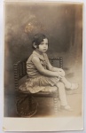 Retrato Infantil de Época  Chancela de Estúdio Carioca (c. 1920-1930)Lindo e melancólico retrato fotográfico original de estúdio, em formato postal vertical. A imagem registra uma menina pequena sentada de perfil sobre uma cadeira infantil de madeira trabalhada em estilo colonial/rústico. Ela veste um clássico vestido plissado ou listrado de época, meias brancas altas e sapatos de boneca, com o cabelo curto ondulado típico da moda infantil daquela geração. O plano de fundo apresenta um sutil degradê pictorialista de estúdio.