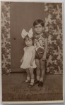 Fotografia Histórica de Estúdio Infantil (1928)  Primos/Irmãos com Inscrição e Data na FrenteRaríssimo e encantador retrato fotográfico de estúdio, original de época, capturado em formato vertical. A imagem registra duas crianças de corpo inteiro posando à frente de um cenário de época decorado com cortinas de padrão floral denso
