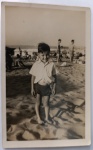 Fotografia Vintage Internacional (1937)  Menino na Praia de South Beach, Durban (África do Sul) por "Scotty"Raríssima e belíssima fotografia vernacular internacional original de época, capturada em plano inteiro vertical com corte reto e fina margem branca. A imagem registra um menino sorridente descalço na areia da praia, vestindo calção de banho escuro e uma camisa social clara de mangas curtas e colarinho aberto.