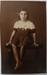Retrato Fotográfico Histórico (1939)  Menina no Estúdio Rogato (Maceió - AL) com DedicatóriaRaríssimo retrato fotográfico de estúdio, original de época, em formato cartão-postal vertical. A imagem registra em plano inteiro uma menina (Vilma) sentada sobre um banco ou banco rústico de estúdio. Ela veste um impecável vestido escuro texturizado ou de poás, destacado por uma ampla e detalhada gola de renda ou crochê clara, além de carregar uma medalha no peito e calçar sandálias de época.