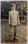 Retrato Fotográfico Vintage  Menino em Terno de Alfaiataria de Época (c. 1920-1930)Belo e expressivo retrato fotográfico original de época, em plano inteiro vertical. A imagem registra um menino posando em um ambiente externo (jardim ou quintal) com vegetação densa ao fundo. Ele veste um elegante e completo terno de alfaiataria claro de corte estruturado, composto por paletó de abotoamento duplo (estilo jaquetão), camisa de colarinho rígido, gravata clara e calças compridas de corte reto, finalizando com sapatos de couro escuros. O cabelo aparece impecavelmente penteado com risca lateral, ditando a estética da moda de sua geração.