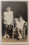 Retrato Fotográfico Vintage de Estúdio  Irmãos/Crianças com Cadeira de Vime (c. 1925-1935)Lindo retrato fotográfico original de estúdio, em formato postal vertical e plano inteiro. A imagem registra duas crianças pequenas posando com trajes infantis leves de verão de cor clara, meias curtas e sapatilhas de fivela claras. A criança à esquerda posa de pé, apoiando a mão no encosto de uma charmosa mini cadeira infantil de vime trançado, onde a segunda criança está sentada de forma solene.