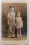   Retrato Fotográfico Histórico de Irmãos / Primos (Niterói, 1930)  Dupla Dedicatória de "Né" e "Nyce"Encantador retrato de estúdio em corpo inteiro de duas crianças pequenas. À esquerda, o menino (Né) posa sorridente vestindo uma autêntica camisa de gola marinheiro clara e calças compridas de alfaiataria de cintura alta com botões aparentes. À direita, a menina (Nyce) veste um delicado vestidinho claro com pregas e babados na barra, meias brancas e sapatilhas. O fundo do estúdio apresenta um suave cenário pictorialista esfumaçado.
