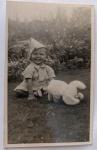 Fotografia Vintage  Bebê com Chapéu de Papel Dobrado e Brinquedo de Pelúcia (c. 1940-1950)Encantadora fotografia original de época, em formato vertical e plano inteiro. A imagem registra um bebê sorridente sentado diretamente sobre a grama em um ambiente externo (jardim ou quintal) com canteiros de flores brancas ao fundo. A criança veste um macaquinho ou conjunto claro de época e calça sapatinhos