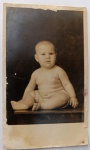 Retrato Fotográfico Histórico de Bebê (1935)  "Luiz Barreto" com Dedicatória de ElísiaClássico retrato de estúdio em corpo inteiro de um bebê gordinho e saudável, vestindo apenas fralda, sentado de perfil e voltado para a frente sobre uma bancada ou mesa de madeira polida. O fundo escuro e infinito do estúdio destaca com excelente contraste a fisionomia e dobras da pele da criança, em uma estética de iluminação muito marcante da década de 1930.