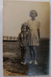   Fotografia Antiga / Gelatina de Prata sobre Papel (Vintage Snapshot). Retrato de Família / Infância ao Ar Livre. Composição com duas meninas em pé em um campo de grama, com uma estrutura de madeira (semelhante a uma ponte ou passarela) ao fundo. A menina menor veste um vestido com suspensórios e um grande laço no cabelo; a mais velha veste um conjunto claro plissado com cinto.