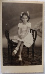  Retrato de Infância / Moda Infantil de Época. Menina sorridente sentada em cadeira clássica de estúdio, com laços simétricos no cabelo e vestido trabalhado de babados. Composição rica com tapete decorado e cenário de fundo pintado. Inscrições no Verso: Carimbo comercial do fotógrafo Carlos W. Weise, detalhando o endereço em Santos e o telefone de quatro dígitos da época ("5784"). Numeração do negativo grafada a lápis.