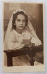 A imagem frontal retrata uma belíssima composição litúrgica de Primeira Comunhão, estimada entre as décadas de 1940 e 1950. A fisionomia da menina se assemelha muito à retratada no lote da jovem "Assunção" (também de Santos), indicando que pode se tratar da mesma pessoa na infância ou de uma irmã próxima. Como o verso não possui dedicatória manuscrita, a identidade nominal exata desta imagem permanece anônima.