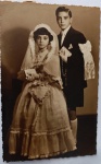 Retrato Religioso / Primeira Comunhão Coletiva / Infância. Composição formal com duas crianças em trajes de comunhão. A menina à esquerda veste um suntuoso vestido longo e rodado com camadas de babados e filó, luvas, véu longo com borda rendada estruturada sobre a cabeça, segurando um missal e um terço. O menino à direita veste um impecável terno escuro com calça curta (bermuda de alfaiataria), meias altas brancas, gravata clara, lenço de bolso e o tradicional laço cerimonial de fita branca com franjas amarrado ao braço esquerdo, também portando missal e terço.