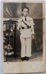  Retrato Religioso / Primeira Comunhão / Identificado. Retrato do menino Ruy em seu dia de comunhão, vestindo o suntuoso uniforme cerimonial branco de estilo militar/gala com dragonas, cinturão e faixa transversal. Composição de estúdio com pedestal de flores e cenário sacro ao fundo. Inscrições no Verso: Rica dedicatória manuscrita utilizando a grafia da época ("offereço", "communhão"), destinada aos seus tios Jovelina e Octavio, assinada por "Ruy", datada e localizada em Santos.