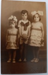  Retrato de Infância / Moda Infantil dos Anos 30 / Identificado. Retrato de estúdio dos três filhos de Dolóres (com forte correlação ao eixo dos irmãos Ary e Odette Oliveira Develly). Ao centro, um menino em pé de bermuda e camisa clara com um grande laço escuro estampado no colarinho, segurando uma bola esférica de brinquedo. Ele é flanqueado por duas meninas (uma menor à esquerda e outra de estatura similar à direita) usando vestidos claros idênticos de babados sobrepostos, cintos escuros e laços volumosos de fita de cetim no cabelo. Inscrições no Verso: Rica dedicatória manuscrita a tinta preta, assinada pela mãe das crianças ("Prima e amiga sincera Dolóres"), direcionada à destinatária Maria. Datada de "4/10/935".