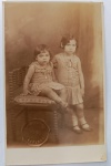   Retrato de Infância / Moda Infantil de Época / Linha Familiar. Retrato formal de duas irmãs em estúdio. A menina menor à esquerda está sentada em uma cadeira de madeira trabalhada com assento estofado em estampa floral, usando um vestido claro com gola redonda (peter pan collar) e medalha em um cordão. A menina maior à direita permanece em pé, com a mão apoiada no encosto da cadeira, vestindo um conjunto claro plissado com cinto fino e cordão com pingente. Ambas usam meias curtas brancas e sapatos boneca escuros. Características Físicas: Fotografia original em tom sépia de excelente definição. Formato cartão-postal clássico com linhas impressas no verso e corte reto tradicional.